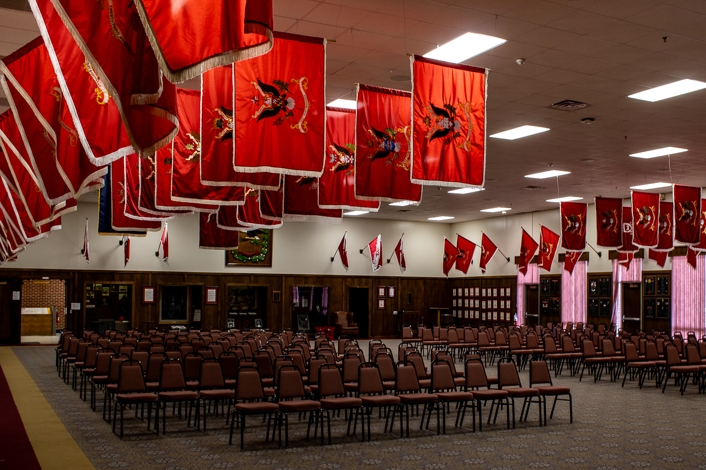 Interior view of the Engineer Regimental Room at the John B. Mahaffey Museum Complex, Fort Leonard Wood with rows of empty chairs facing a display of red Engineer Unit Colors, hanging from the ceiling and walls. The flags create a formal, patriotic atmosphere, complemented by soft lighting and a structured seating arrangement. The walls display plaques and framed memorabilia, showcasing the hall’s dedication to honoring military history and tradition.