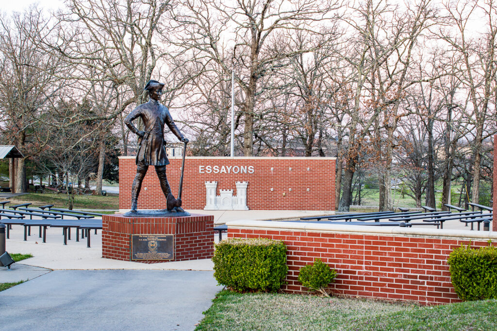 Outdoor statue of a historical U.S. Army Engineer with an axe, standing in front of the 'Essayons' brick memorial wall. The scene includes benches for visitors, well-maintained landscaping, and a backdrop of leafless trees, highlighting the solemn atmosphere and tribute to U.S. Army Engineers