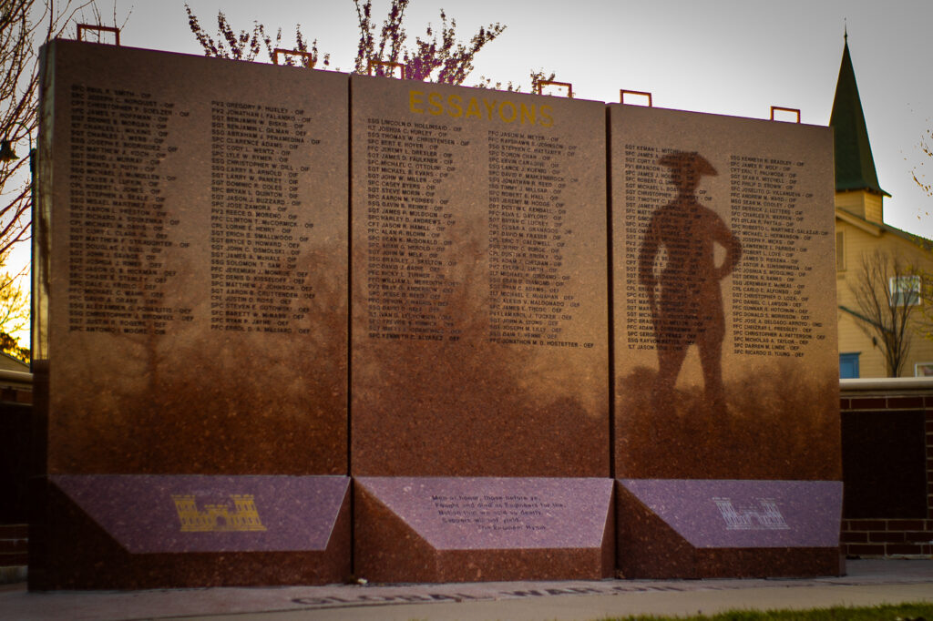 Close-up view of the Essayons Memorial Wall, engraved with names of fallen U.S. Army Engineers. The wall is composed of polished stone panels, with gold 'Essayons' lettering at the top and the symbolic Engineer Castle insignia at the bottom. The shadow of a historical engineer figure is visible on the right panel, symbolizing respect and remembrance. A nearby church steeple and trees provide a serene backdrop, adding to the solemn ambiance.